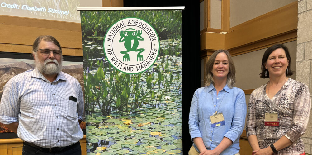 A photo of WWA's Tracy Hames (far left), NAWM's Marla Stelk (middle), and WWA's Erin O'Brien posing with a banner for the NAWM at the Annual State/Tribal/Federal Coordination Meeting hosted by the National Association of Wetland Managers (NAWM).