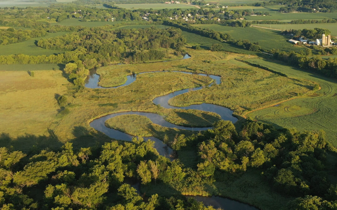 Aerial view of rural landscape with lush greenery, trees, river and wetlands in the foreground and a farmhouse, farmland in the background.